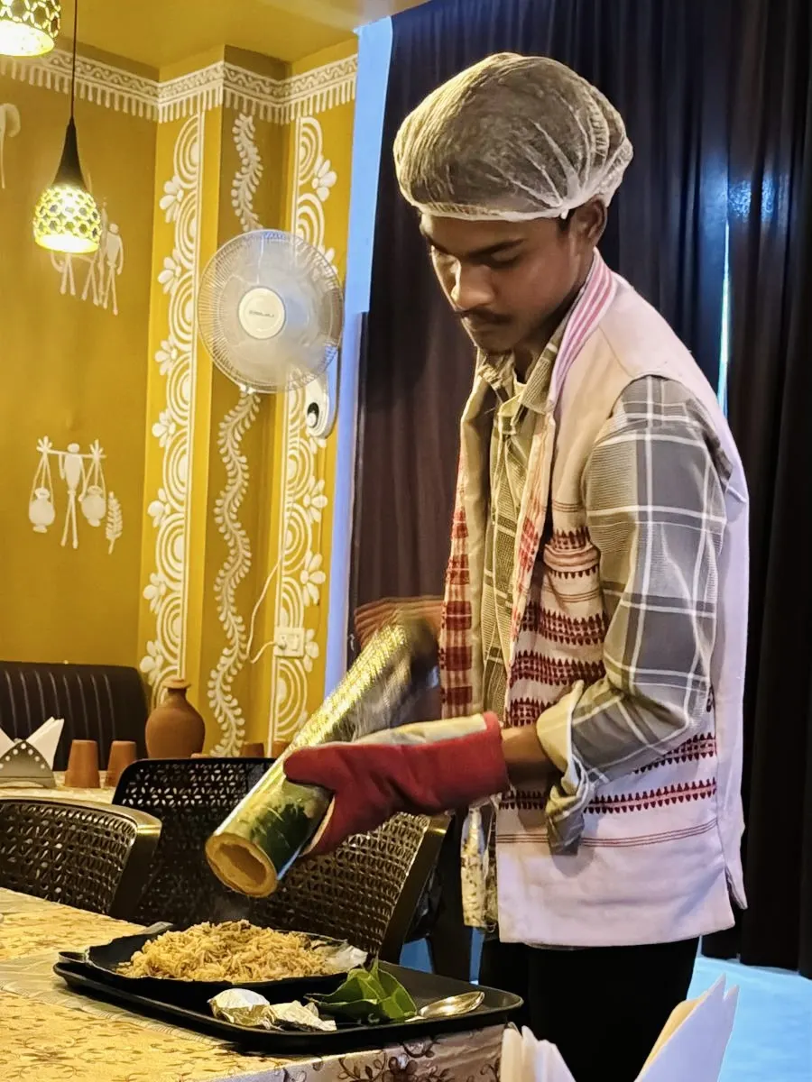 Staff member in traditional tribal attire serving authentic tribal food at JOMHA